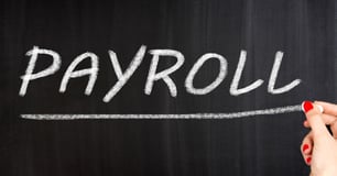 Hand with red nail polish writes Payroll and underlines it with chalk on a chalkboard
