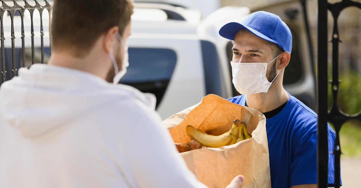 Package delivery person wearing a mask hands off a brown bag of groceries to a man